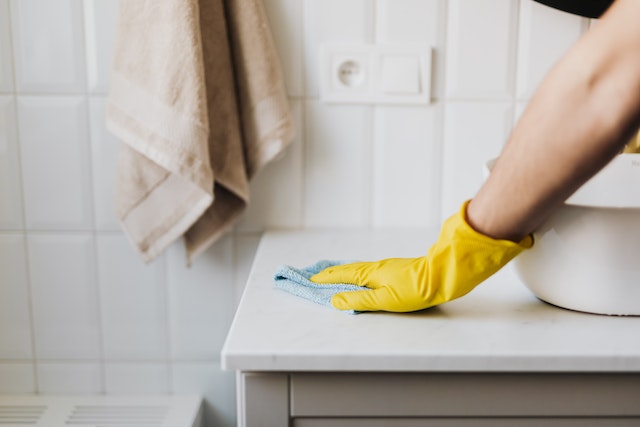 Hand wearing a yellow rubber glove wiping down bathroom sink with a blue washcloth