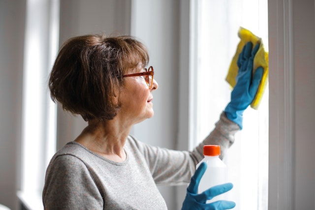 Older person with short brown hair wearing blur rubber gloves and wiping a window with a yellow washcloth