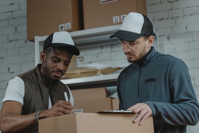 Two people wearing caps looking at a document in room full of mail and packages