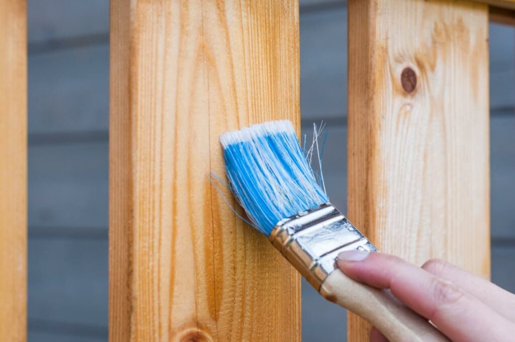 Hand holding a paintbrush against a brown wooden fence
